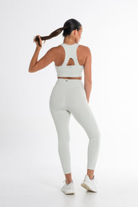 Back view of a woman wearing a matching mint green activewear set, featuring high-waisted leggings and a racerback sports bra with a small keyhole detail. She holds her ponytail with one hand, showcasing the comfortable fit and stylish design. The outfit is paired with white athletic sneakers, and the background is a clean, minimalist white.
