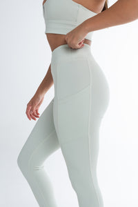 Side view of a woman wearing a light mint ice activewear set, including high-waisted leggings with side pockets and a matching sports bra. She is adjusting the waistband of the leggings, showcasing the smooth, supportive fit. The background is minimal and white, highlighting the sleek, seamless design of the activewear.