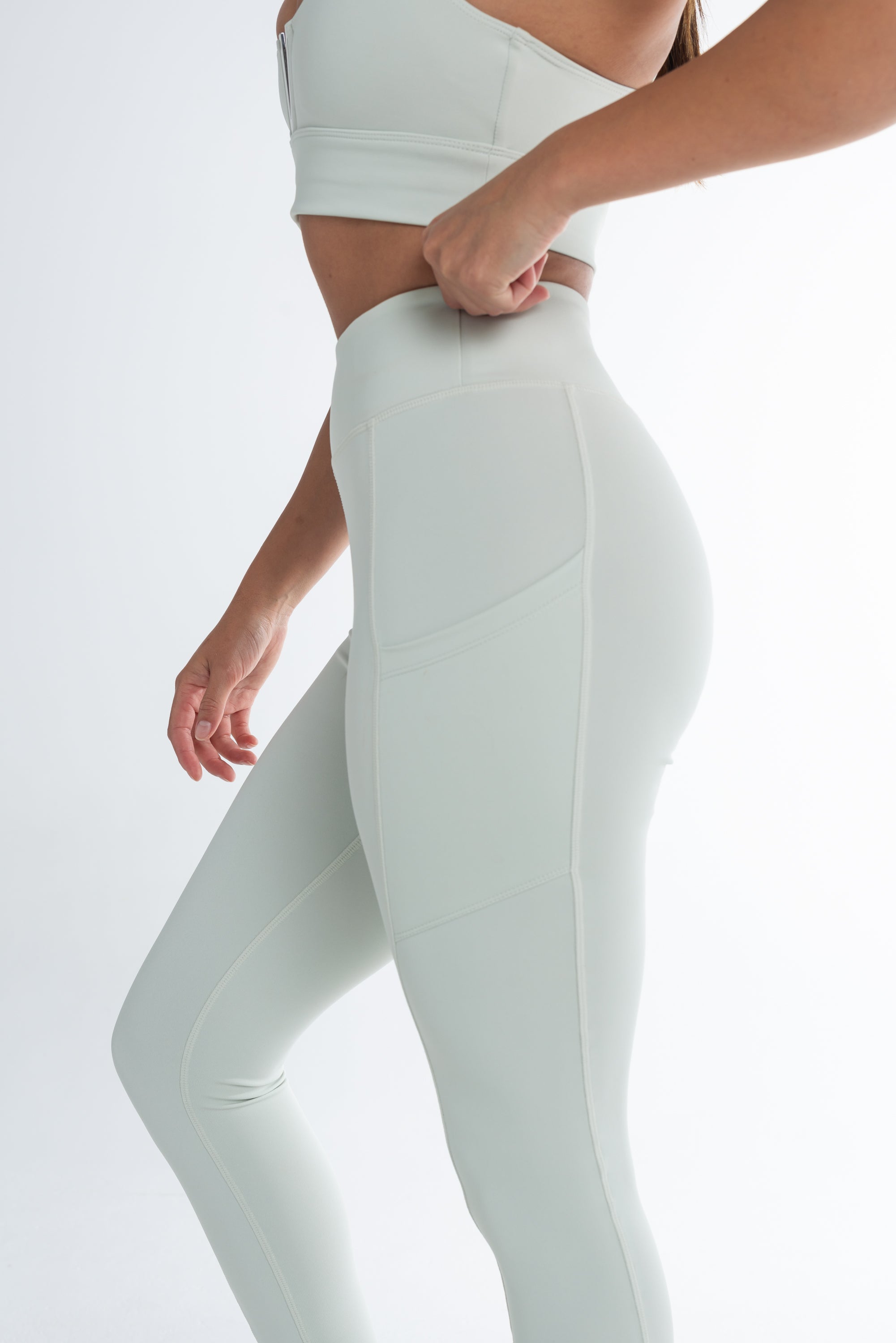 Side view of a woman wearing a light mint ice activewear set, including high-waisted leggings with side pockets and a matching sports bra. She is adjusting the waistband of the leggings, showcasing the smooth, supportive fit. The background is minimal and white, highlighting the sleek, seamless design of the activewear.