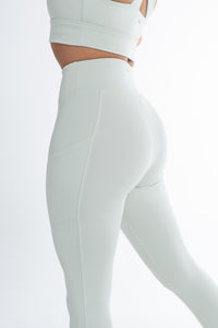 Back view of a woman wearing a matching light mint ice activewear set, including a high-waisted leggings and a fitted sports bra. The leggings feature side pockets and a smooth, contouring design that enhances the body’s natural shape. The background is a clean, minimalistic white, highlighting the sleek, seamless look of the activewear.