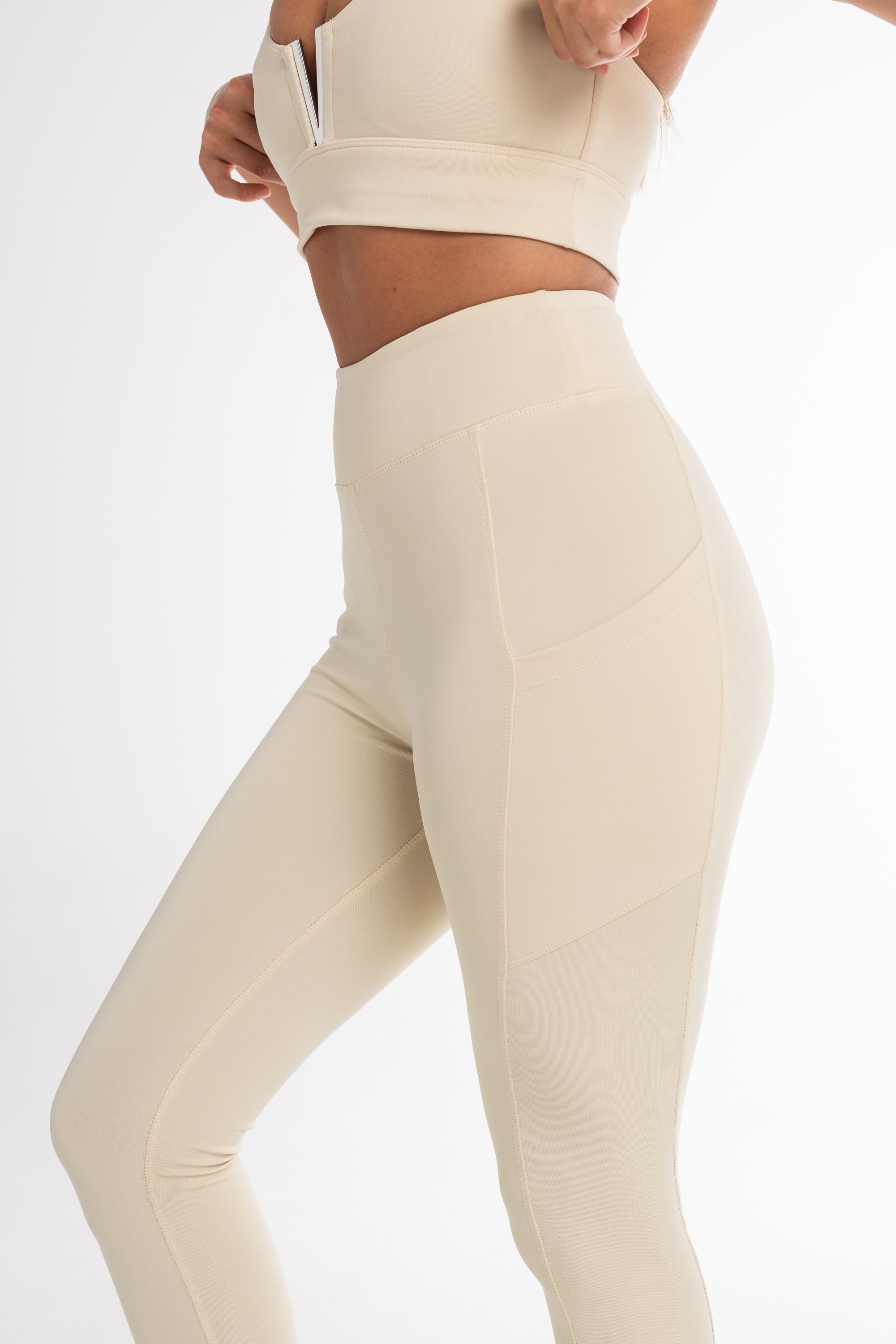 Close-up of a woman wearing a cream sports bra and high-waisted leggings, showcasing a sculpted fit with side pockets and a seamless, breathable design. Ideal for active and versatile wear.