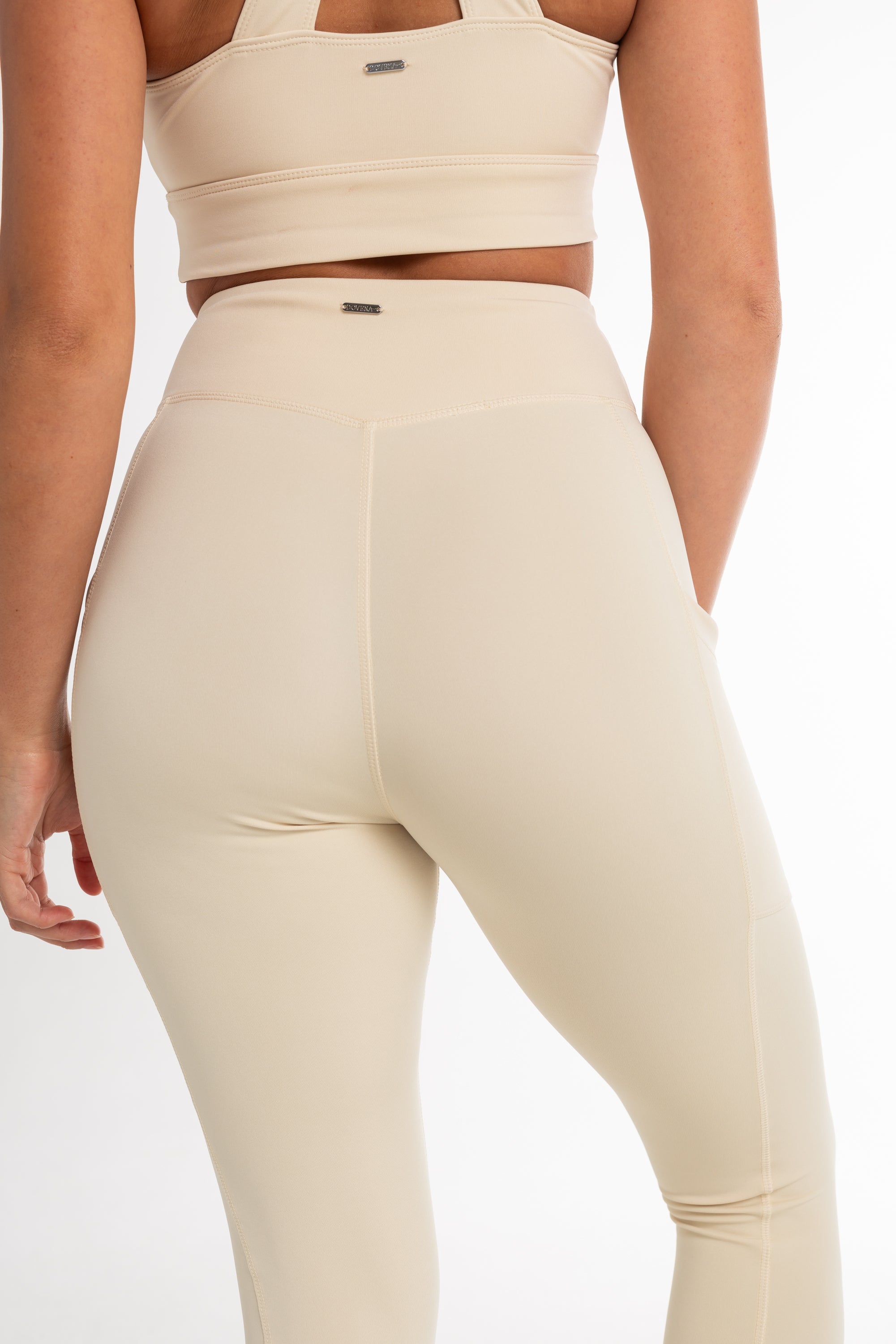 Back view of a woman wearing a beige sports bra and high-waisted leggings with a sculpted fit. The set features a minimalist design and seamless fabric for comfortable movement.