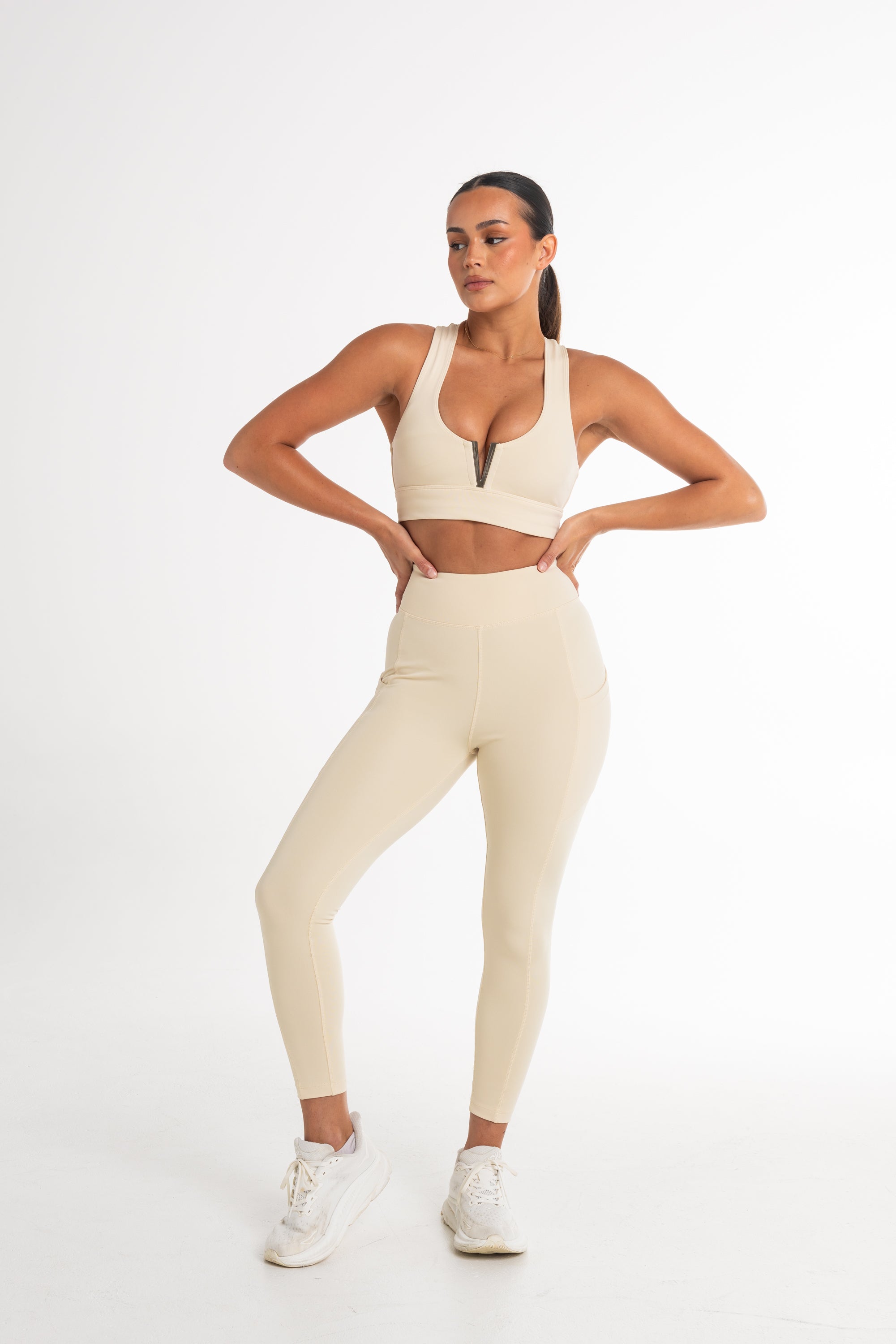 Full-body shot of a woman in a cream sports bra and high-waisted leggings. The activewear set highlights a sleek, body-contouring fit with side pockets, perfect for versatile and stylish movement.