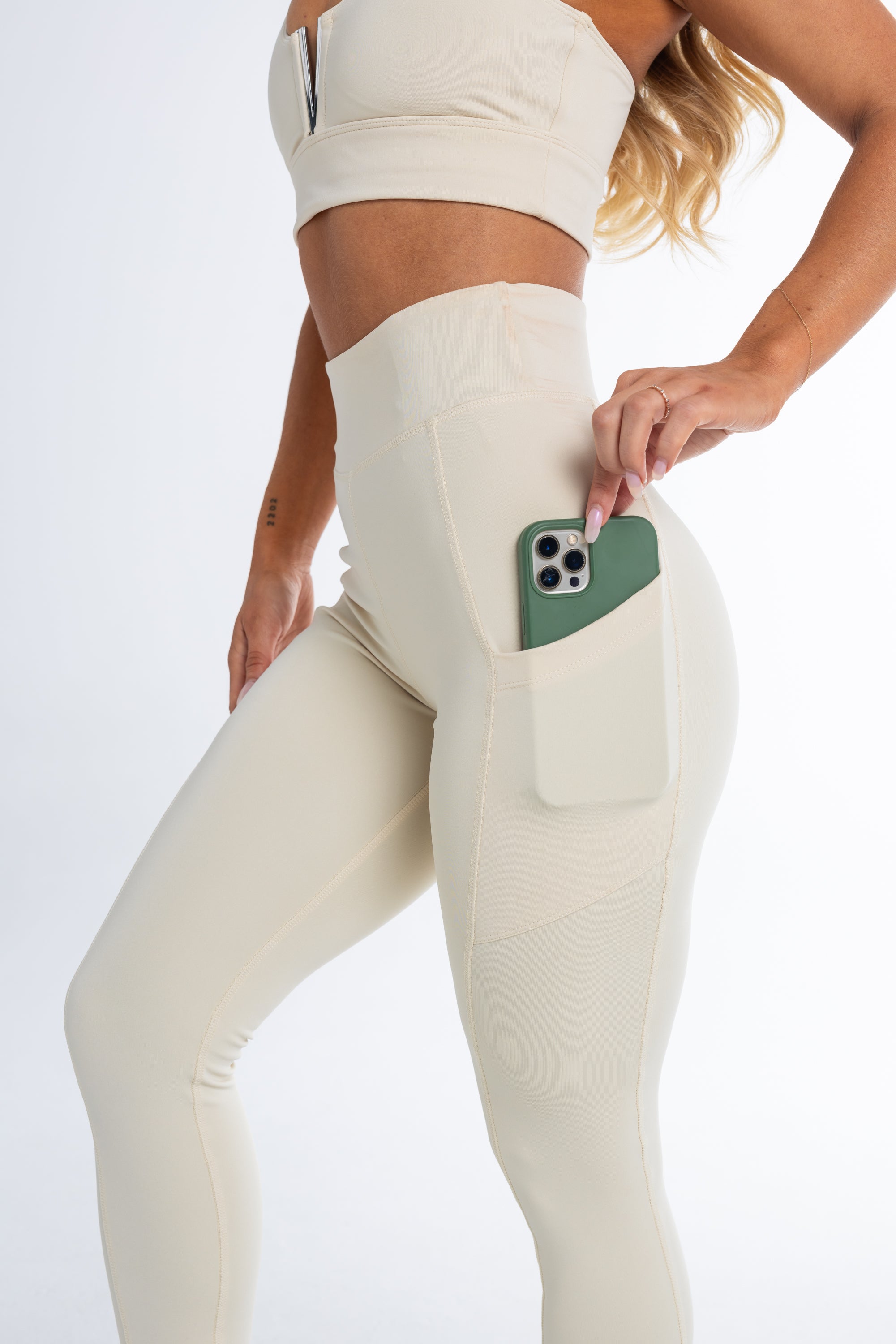 Full length image of young woman wearing a matching sports bra and sculpting activewear 7/8 leggings in cream with side pockets showing a phone sliding into pockets